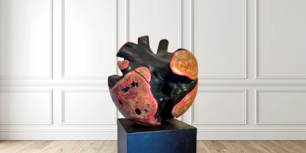Abstract wood sculpture with textured red and black surfaces displayed on a pedestal in a gallery-style interior.